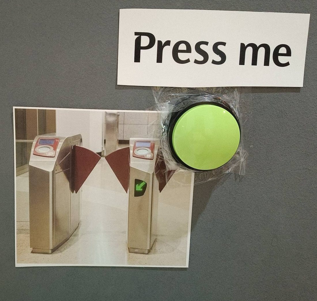A printed photo of MRT fare gates on the wall, with a big green 'Press me' button next to it